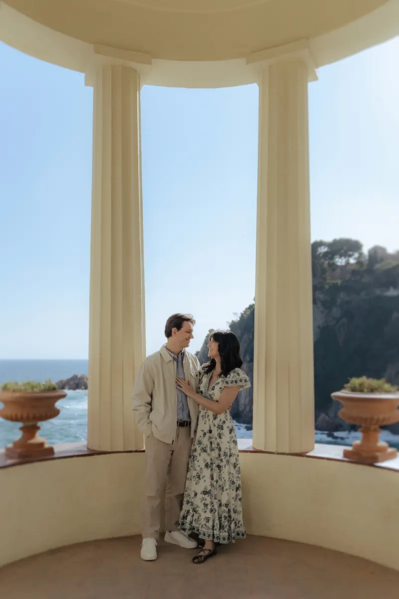 Couple gazes at each other, smiling beneath seaside columns during a surprise proposal photoshoot.