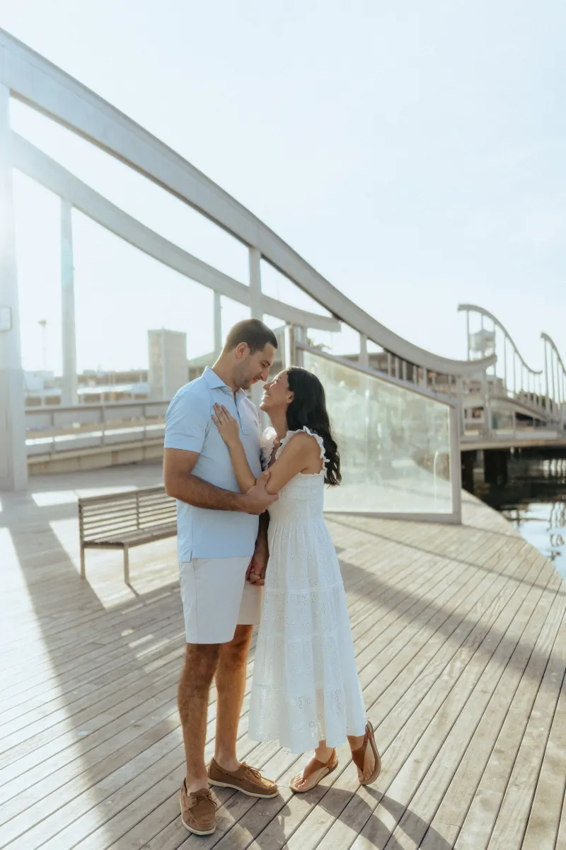Couple gazes lovingly at each other on a sunlit marina boardwalk during a couples & engagements photoshoot session.