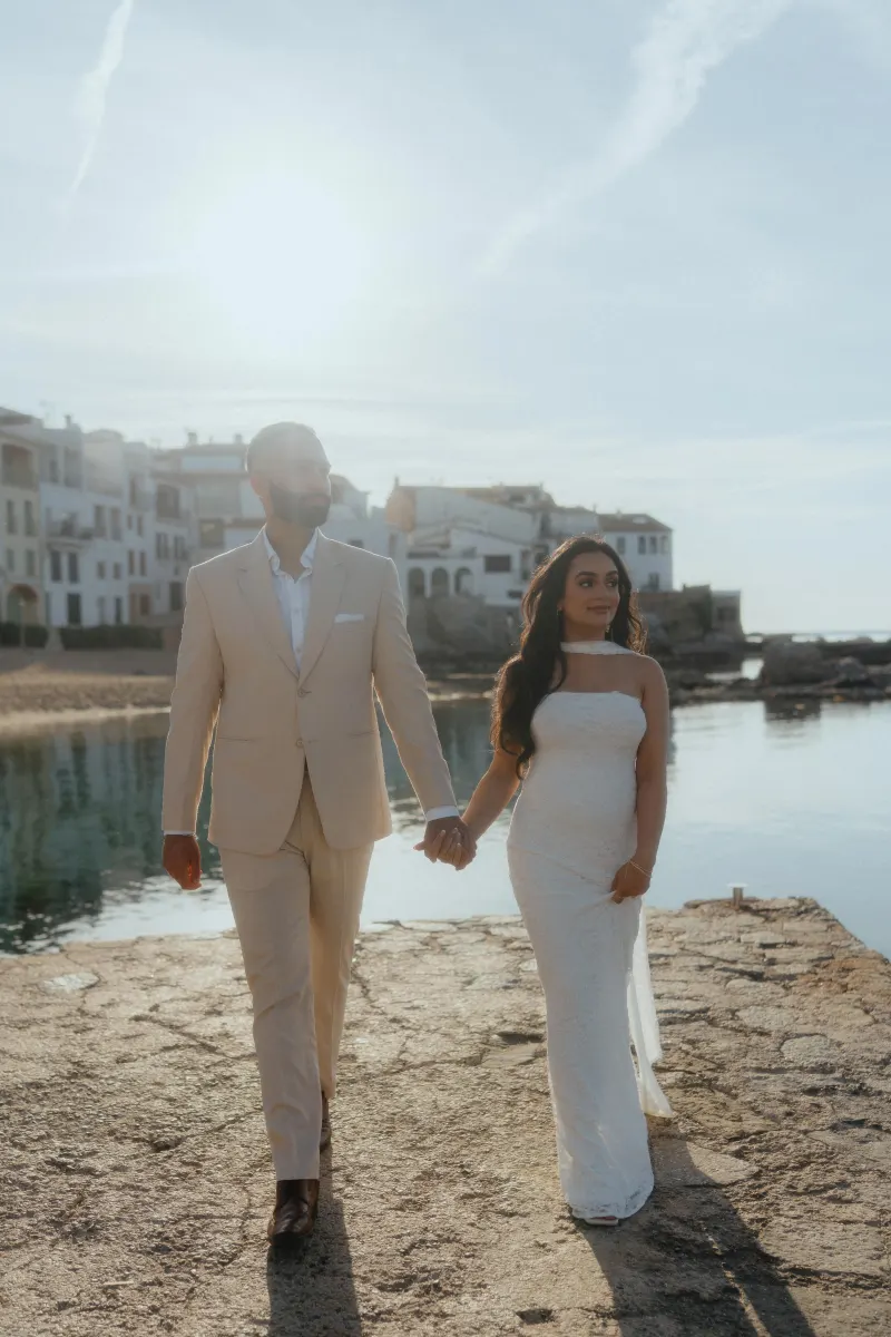 Couple holding hands after a surprise proposal photoshoot, walking by the water in soft golden sunlight.