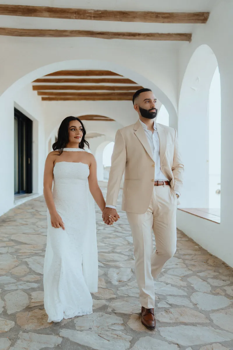 Couple holding hands after a surprise proposal photoshoot, walking through a sunlit Mediterranean-style corridor.