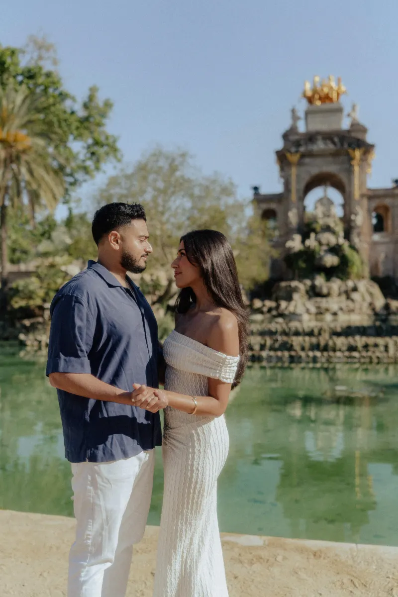 Couple holding hands and gazing at each other by a scenic fountain during a romantic engagement photoshoot outdoors.