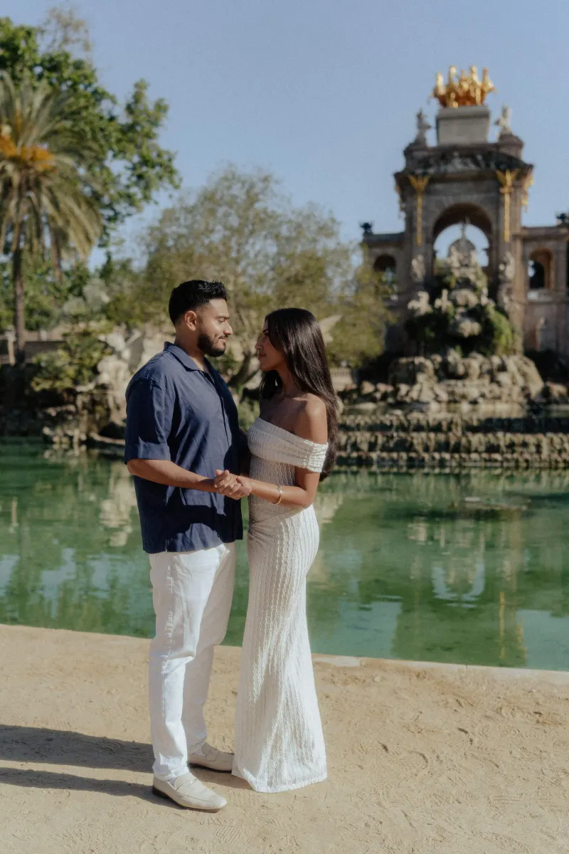 Couple holding hands and gazing at each other by a sunlit fountain during a romantic engagement photoshoot.