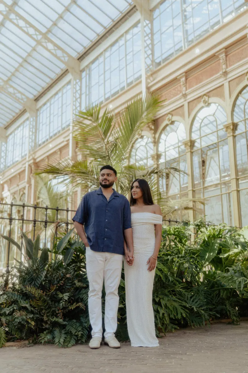Couple holding hands and sharing a quiet moment during an engagement session in a sunlit glasshouse.