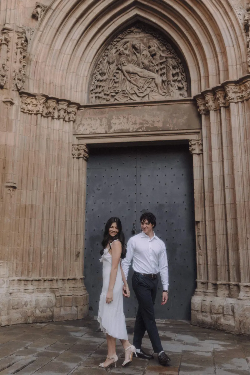 Couple holding hands and smiling as they walk past a historic cathedral door during an engagement photoshoot.