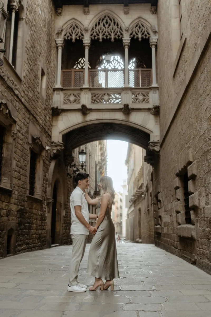 Couple holding hands and smiling at each other beneath Barcelona’s Gothic bridge during a Vacation Portraits photoshoot.