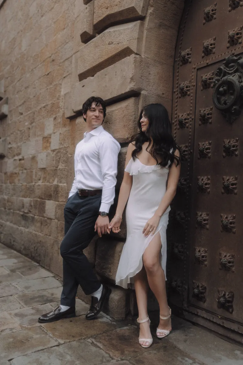 Couple holding hands and smiling at each other by a rustic doorway during a romantic engagement photoshoot in the city.