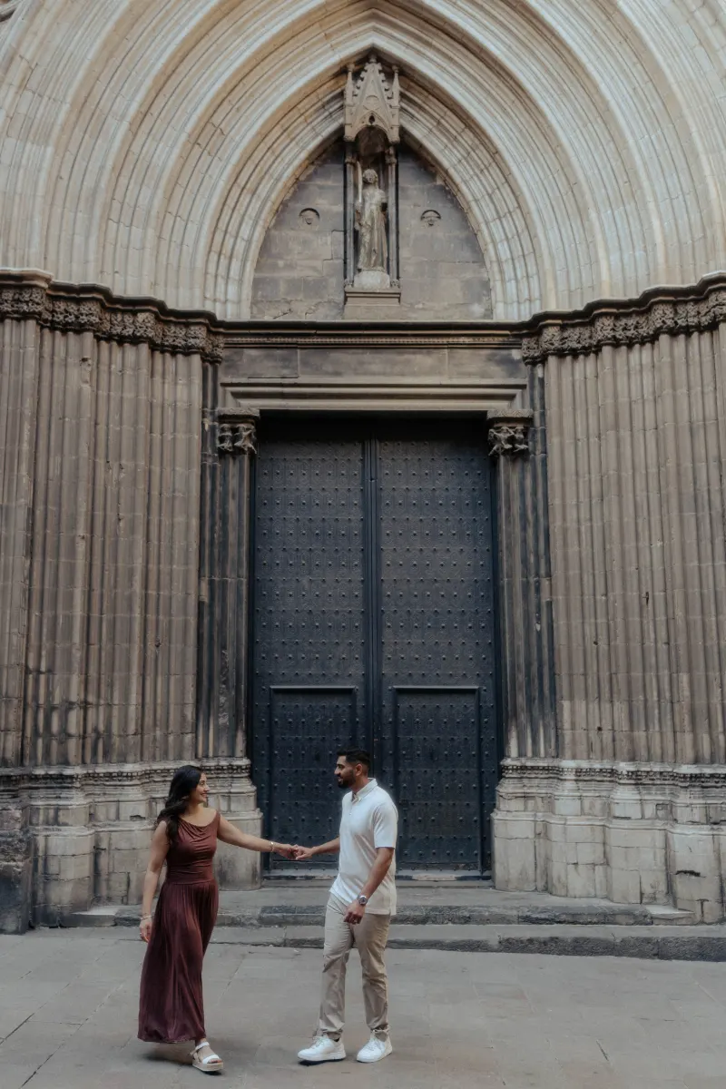 Couple holding hands and smiling at each other in front of a grand gothic doorway during their engagement photoshoot.