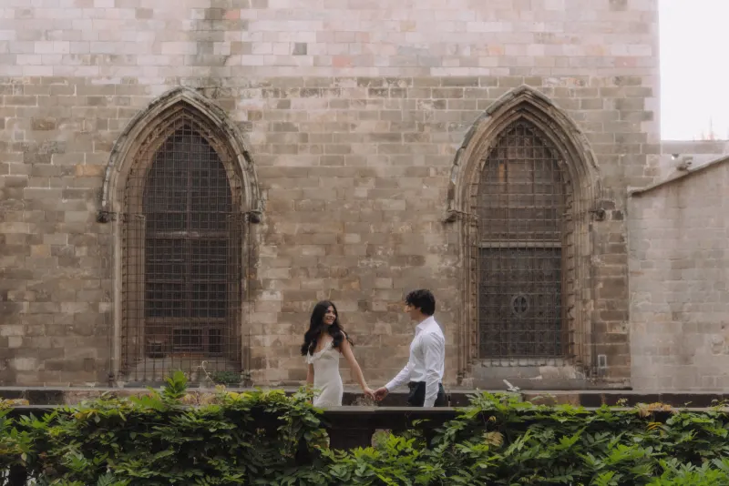 Couple holding hands and smiling at each other in front of a historic stone building during an engagement photoshoot.