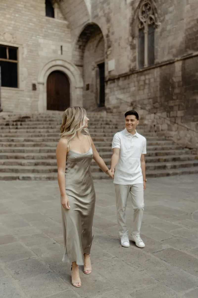 Couple holding hands and smiling at each other in front of historic stone steps during a Vacation Portraits shoot in Barcelon