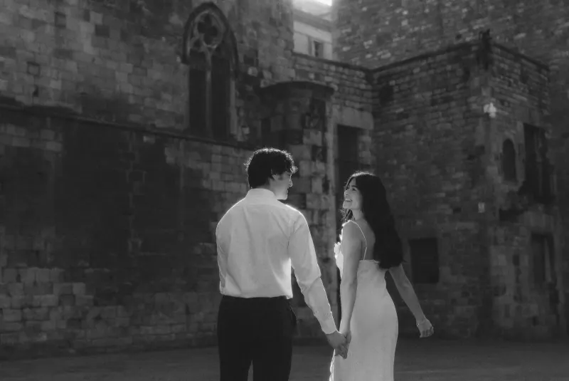 Couple holding hands and smiling at each other in soft light during a romantic engagement photoshoot by historic stone walls.
