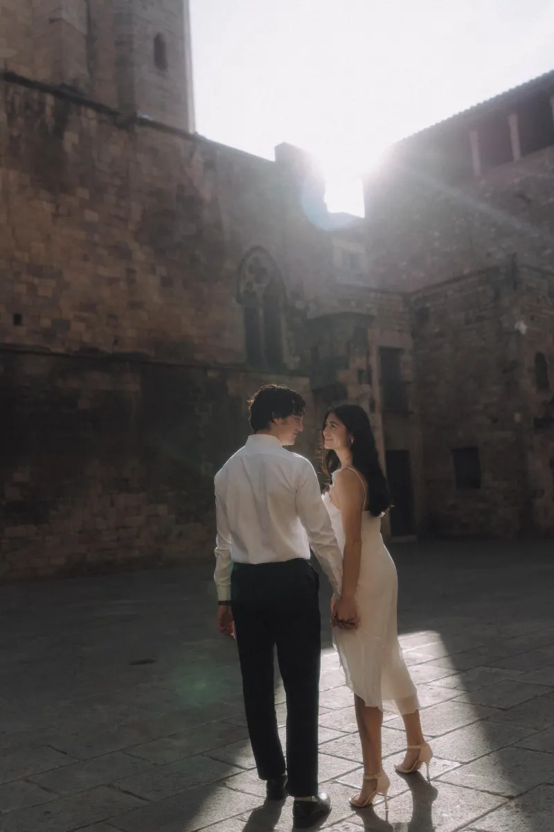Couple holding hands and smiling at each other in soft sunlight during a romantic engagement photoshoot in the Gothic Quarter