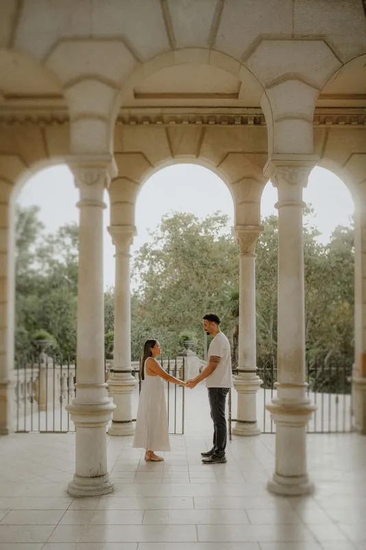 Couple holding hands and smiling at each other under stone arches during a romantic engagement photoshoot in the park.