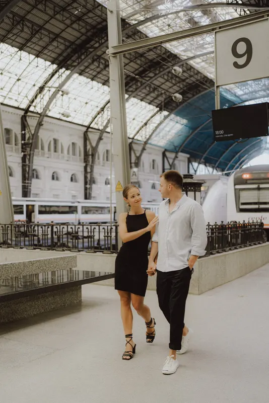 Couple holding hands and smiling at each other while walking through a sunlit train station during an engagement photoshoot.