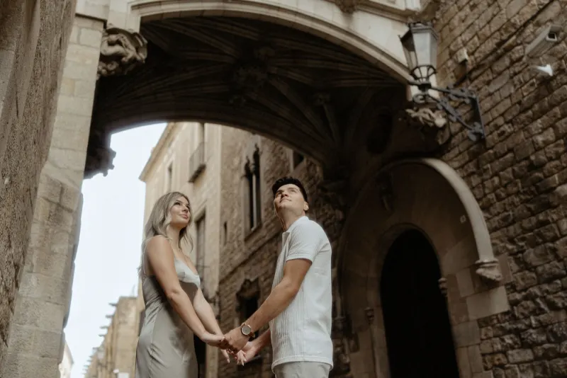 Couple holding hands and smiling beneath a historic archway in Barcelona during a Vacation Portraits photoshoot.