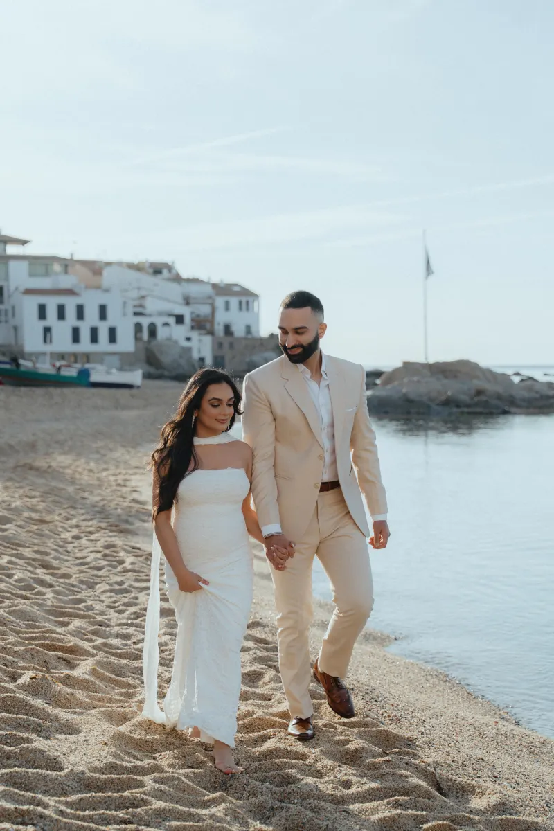 Couple holding hands and smiling during a surprise proposal photoshoot on a sunny beach in a coastal village.