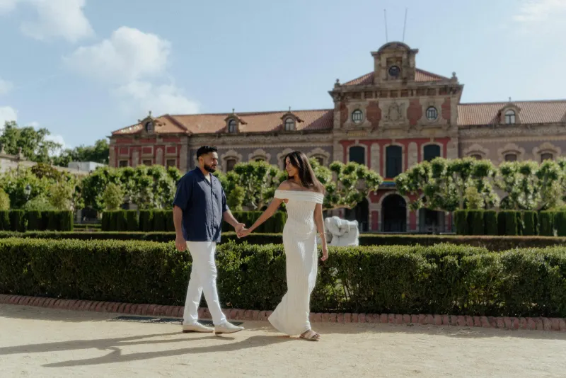 Couple holding hands and strolling through a sunlit garden in Barcelona during a romantic engagement photoshoot.