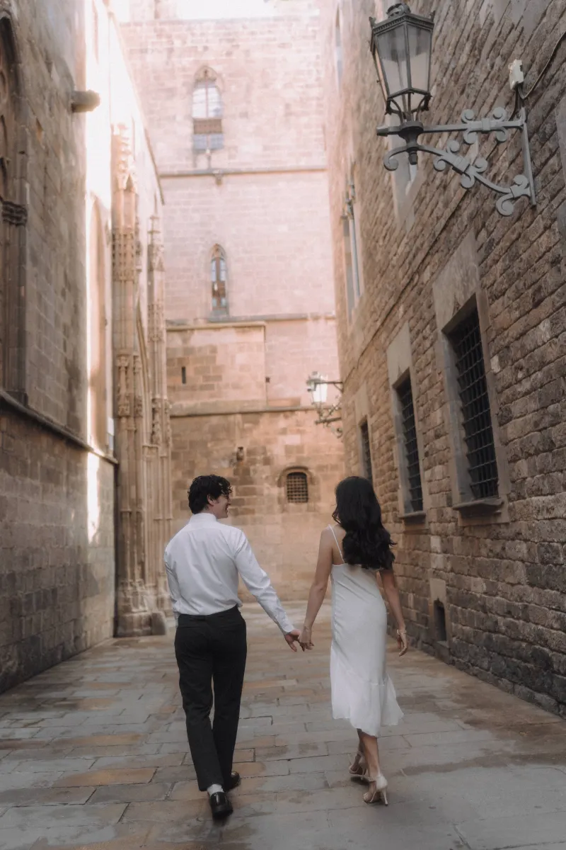Couple holding hands and strolling through a sunlit Gothic Quarter alley during a romantic couples & engagements photoshoot.