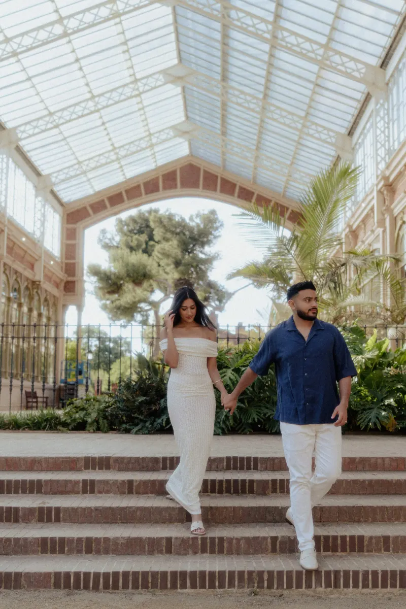 Couple holding hands and walking down steps in a sunlit conservatory during a relaxed engagement photoshoot.