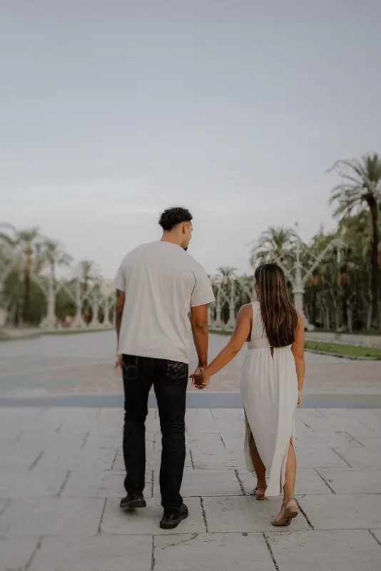 Couple holding hands and walking together at sunset, captured during an engagement photoshoot in a palm-lined park.