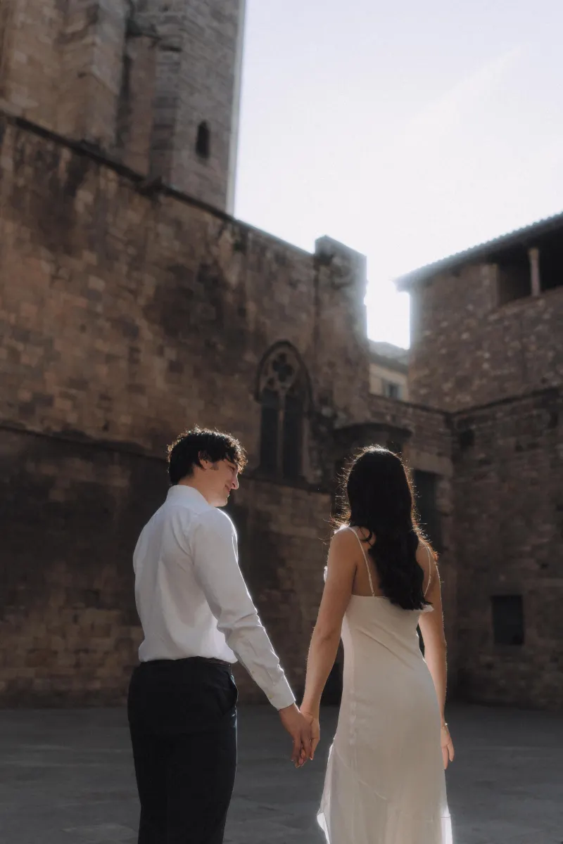 Couple holding hands and walking together in soft sunlight, enjoying a romantic engagement photoshoot in a historic courtyard