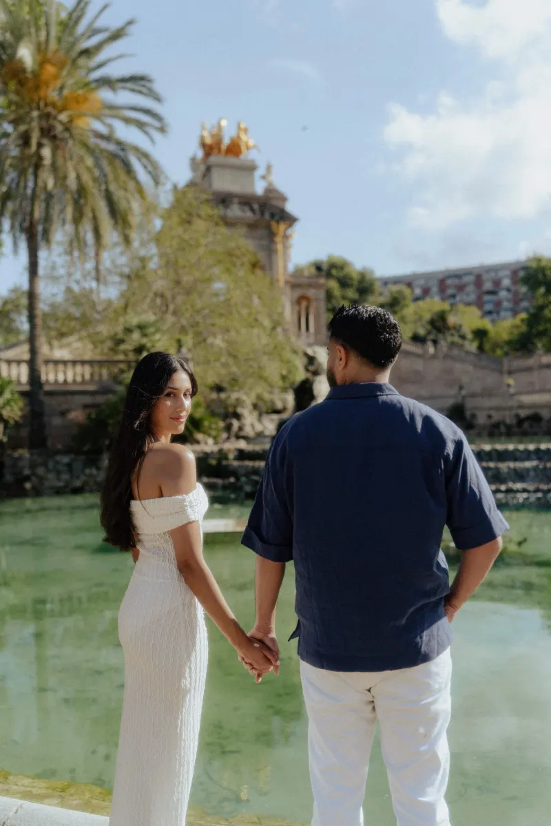 Couple holding hands by a tranquil pond, sharing a soft glance during a romantic engagement photoshoot outdoors.