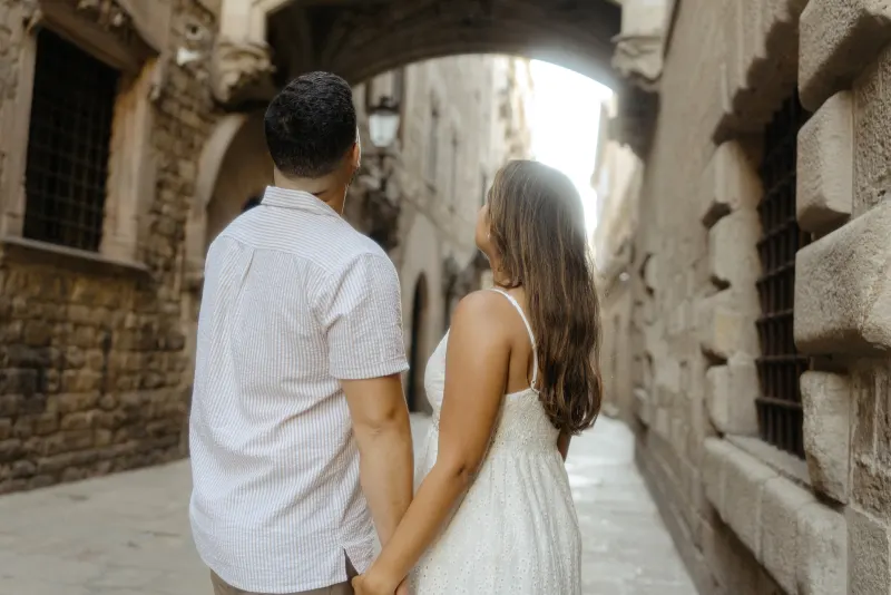 Couple holds hands and gazes up at historic stone archway during a romantic engagement photoshoot in the old city.