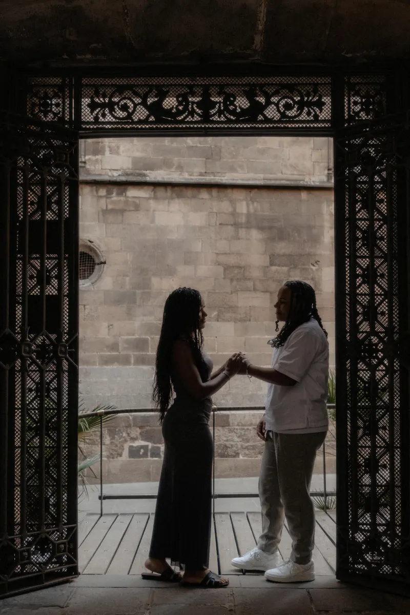 Couple holds hands and shares a heartfelt moment beneath ornate gates during a surprise proposal photoshoot in Barcelona.