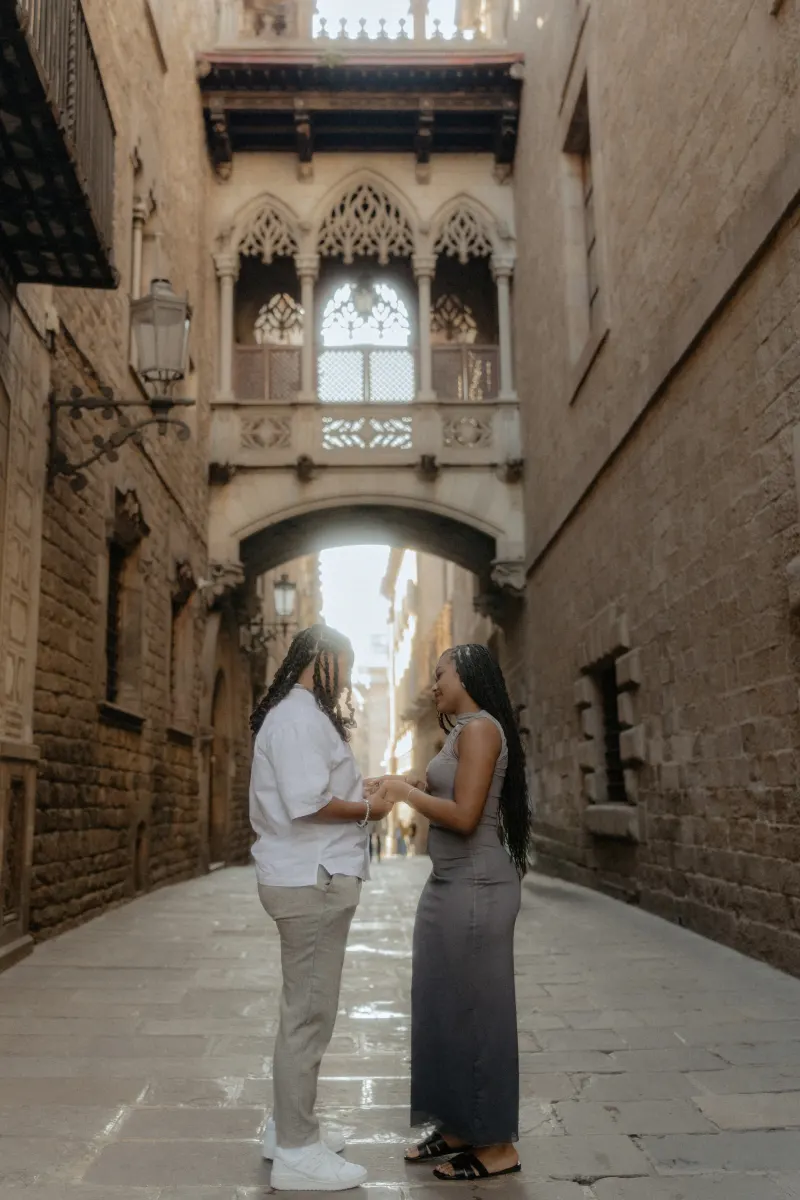 Couple holds hands and smiles at each other beneath Barcelona’s Gothic bridge during a surprise proposal photoshoot.