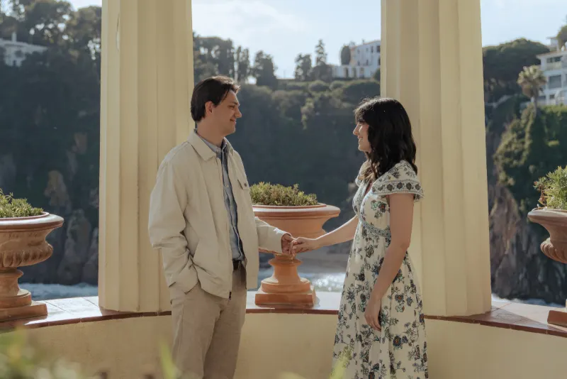 Couple holds hands and smiles at each other under sunlit columns during a surprise proposal photography session by the sea.