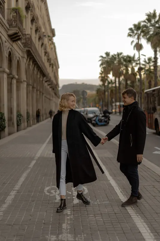 Couple holds hands and smiles at each other while strolling a palm-lined street in Barcelona during an engagement session.