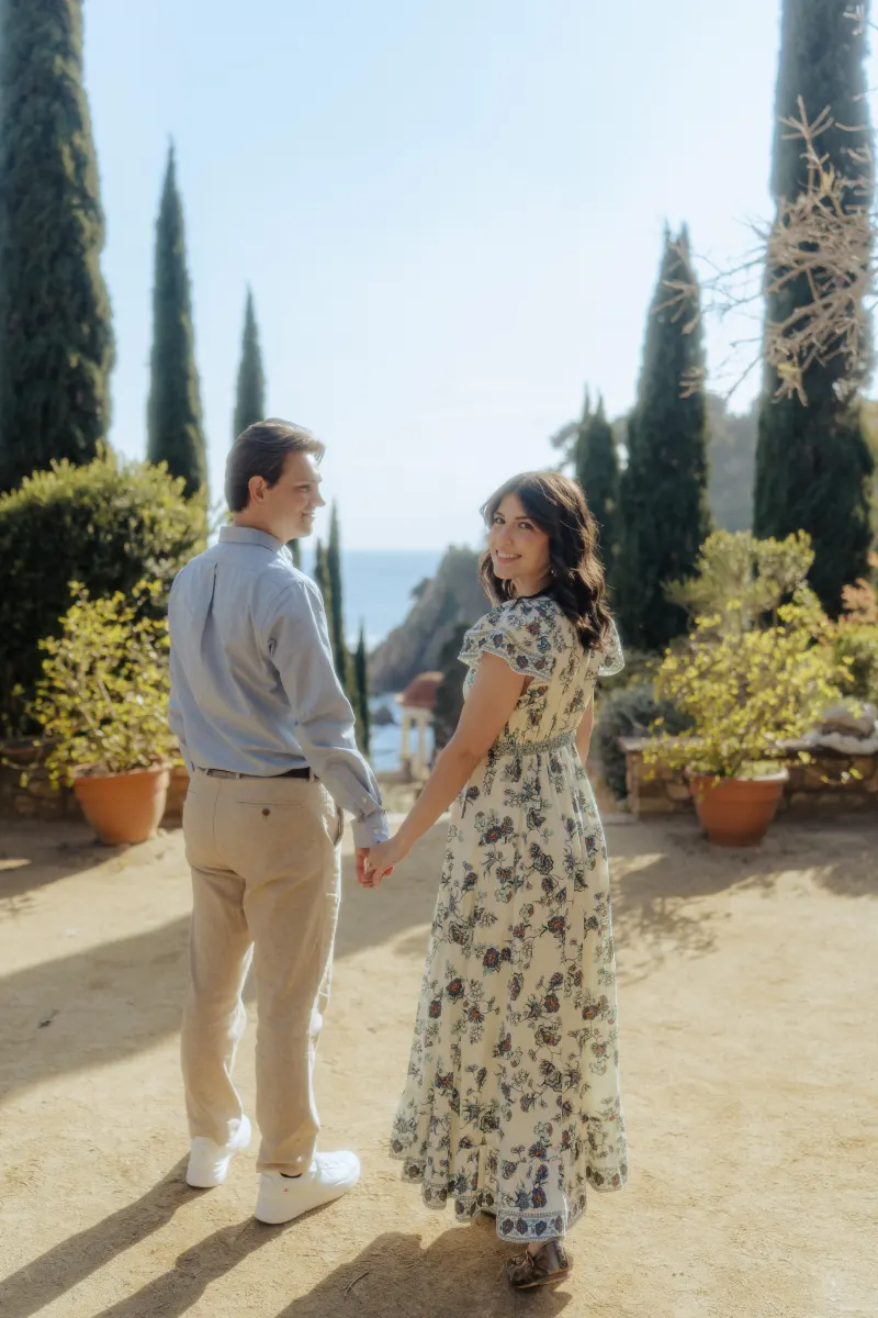 Couple holds hands and smiles in a Mediterranean garden, celebrating their engagement during a surprise proposal photoshoot.