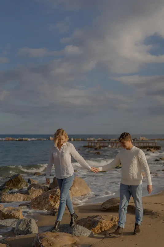 Couple holds hands as they walk over rocks by the sea during a couples & engagements photoshoot in Barcelona.