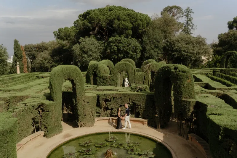 Couple holds hands by a fountain in a manicured garden maze during a surprise proposal photoshoot.