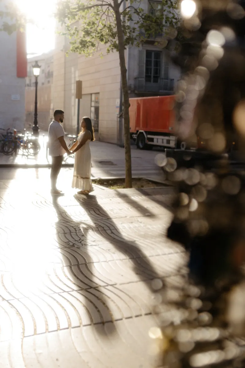 Couple holds hands in soft sunlight on a city street, sharing a quiet moment during their engagement photoshoot.