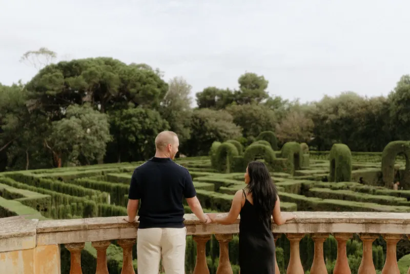 Couple holds hands on a stone terrace, sharing a quiet moment overlooking a garden maze during a surprise proposal photoshoot