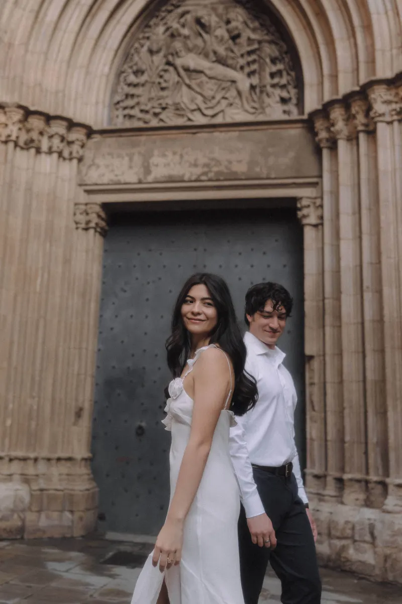 Couple in elegant attire walks hand in hand, sharing a soft smile during a couples & engagements photoshoot by a cathedral.