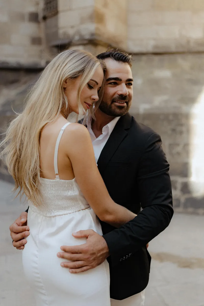 Couple in elegant outfits share a warm embrace on a sunlit city street during a couples & engagements photoshoot.