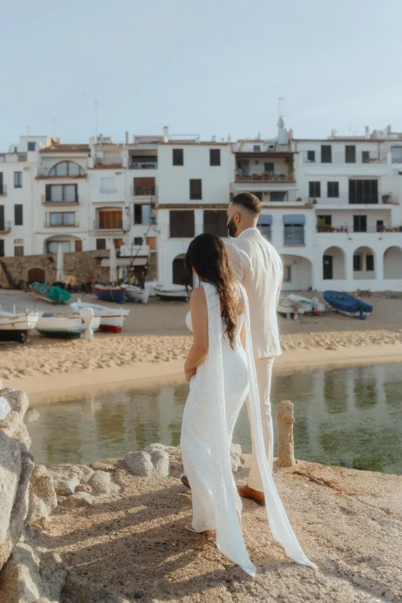 Couple in elegant white outfits share a quiet moment by the water during a surprise proposal photoshoot in a coastal village.