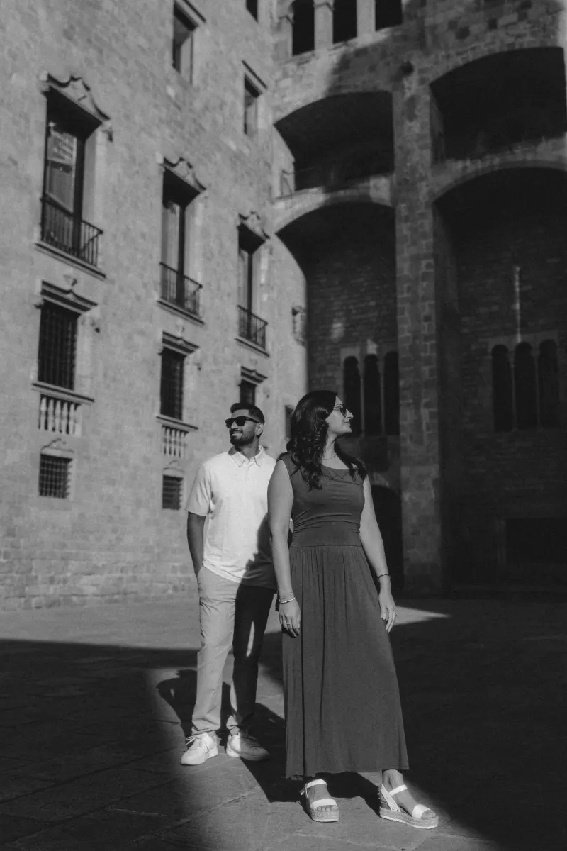 Couple in stylish summer outfits shares a relaxed moment during an engagement photoshoot in a sunlit historic courtyard.