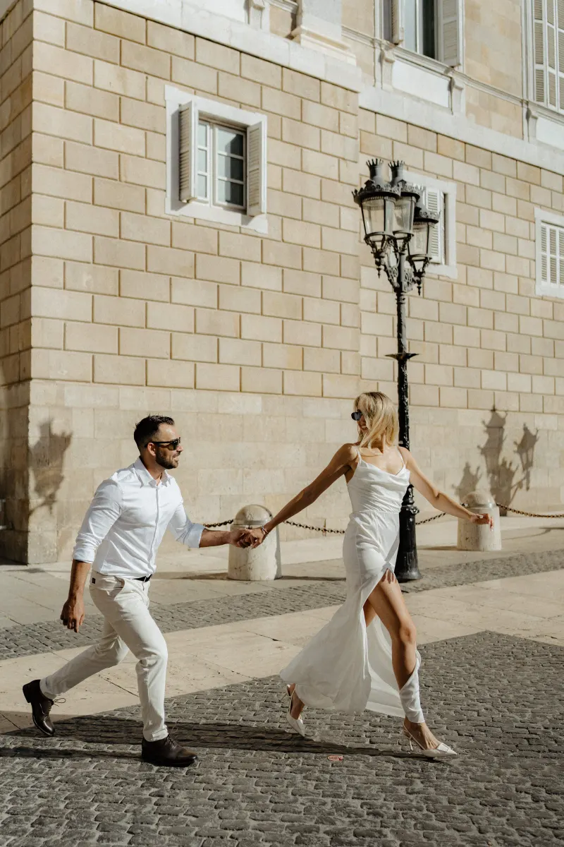 Couple in white outfits laughing and holding hands as they stroll through a sunlit city street during an engagement session.