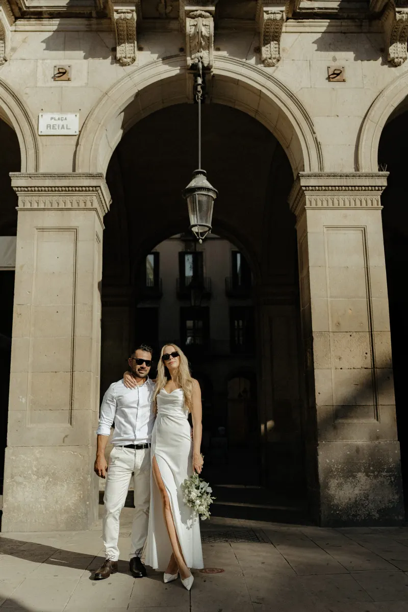 Couple in white outfits pose together under historic city arches, celebrating their engagement in natural sunlight.