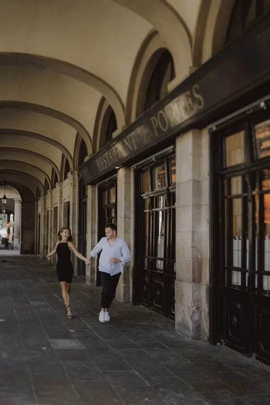 Couple laughing and holding hands as they stroll under elegant arches during a couples & engagements photoshoot.