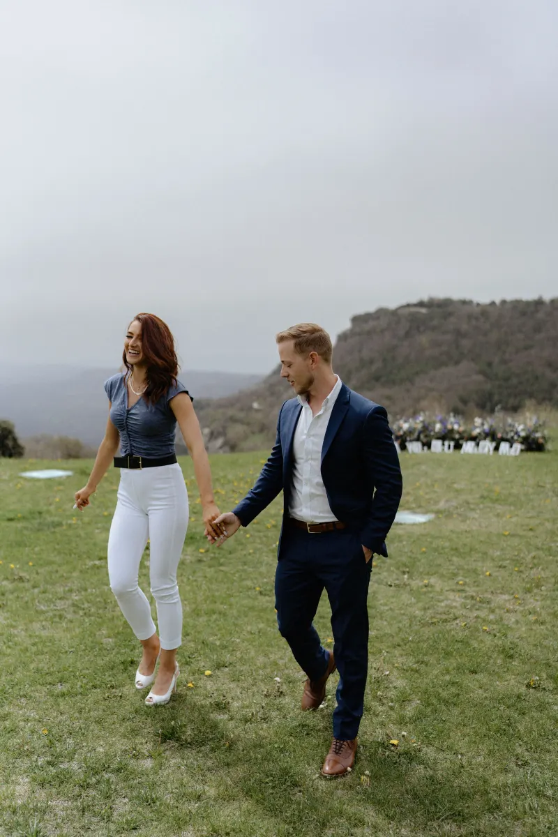 Couple laughing and holding hands while walking on a scenic hillside during a surprise proposal photoshoot.