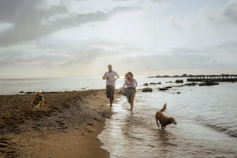 Couple laughing as they run barefoot along the beach with their dogs, captured during a couples & engagements session.