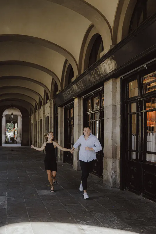 Couple laughing as they run hand in hand beneath elegant arches during a couples & engagements photoshoot in the city.