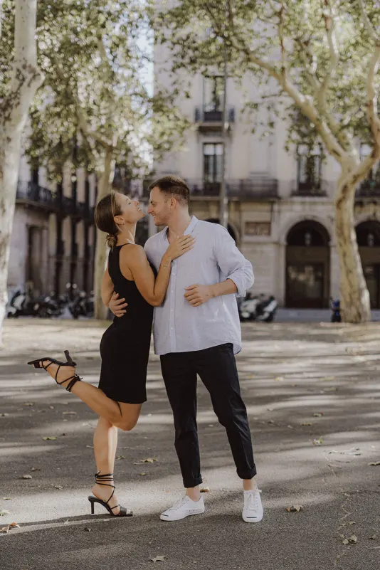 Couple laughing together under leafy trees in the city, enjoying a lighthearted moment during an engagement photoshoot.