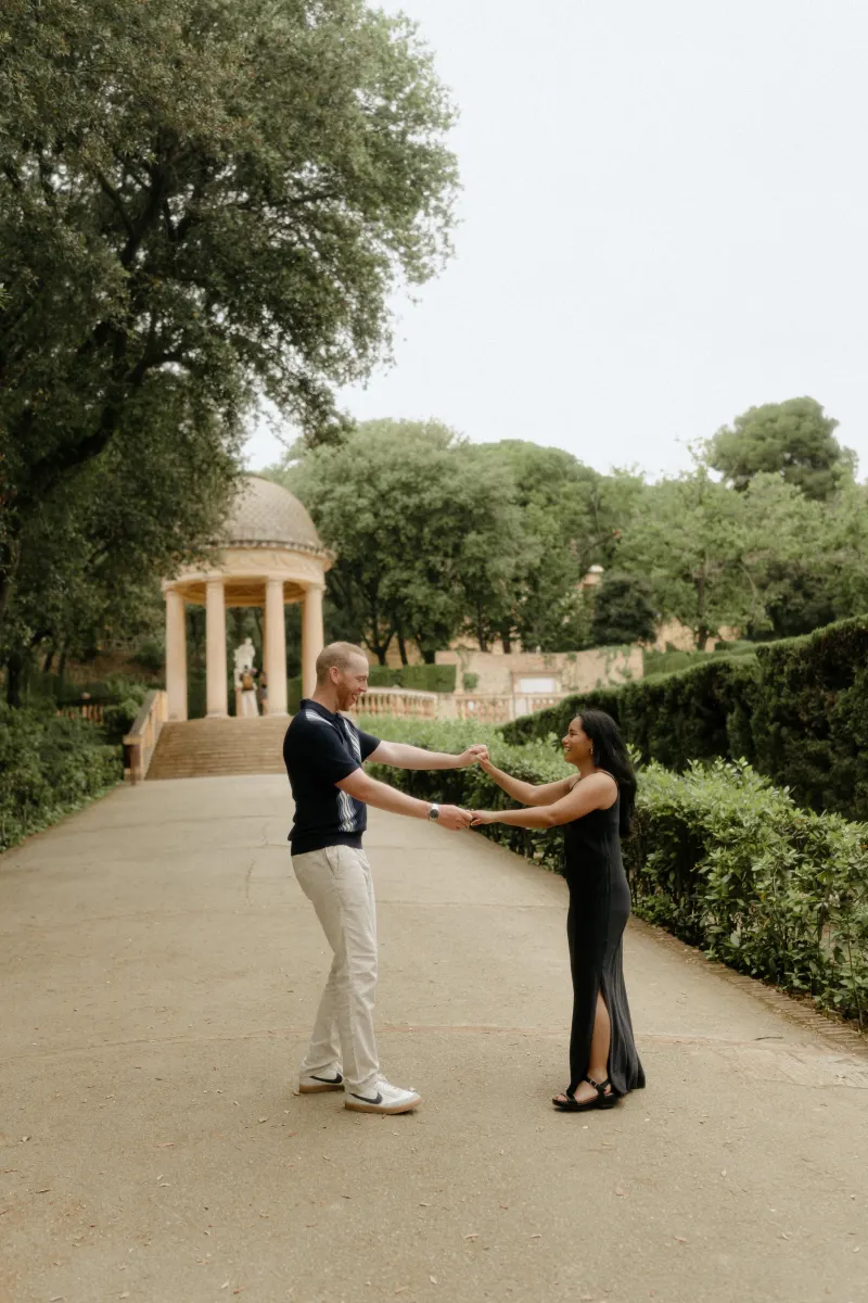 Couple laughs and holds hands on a garden path, sharing a joyful moment during a surprise proposal photoshoot.