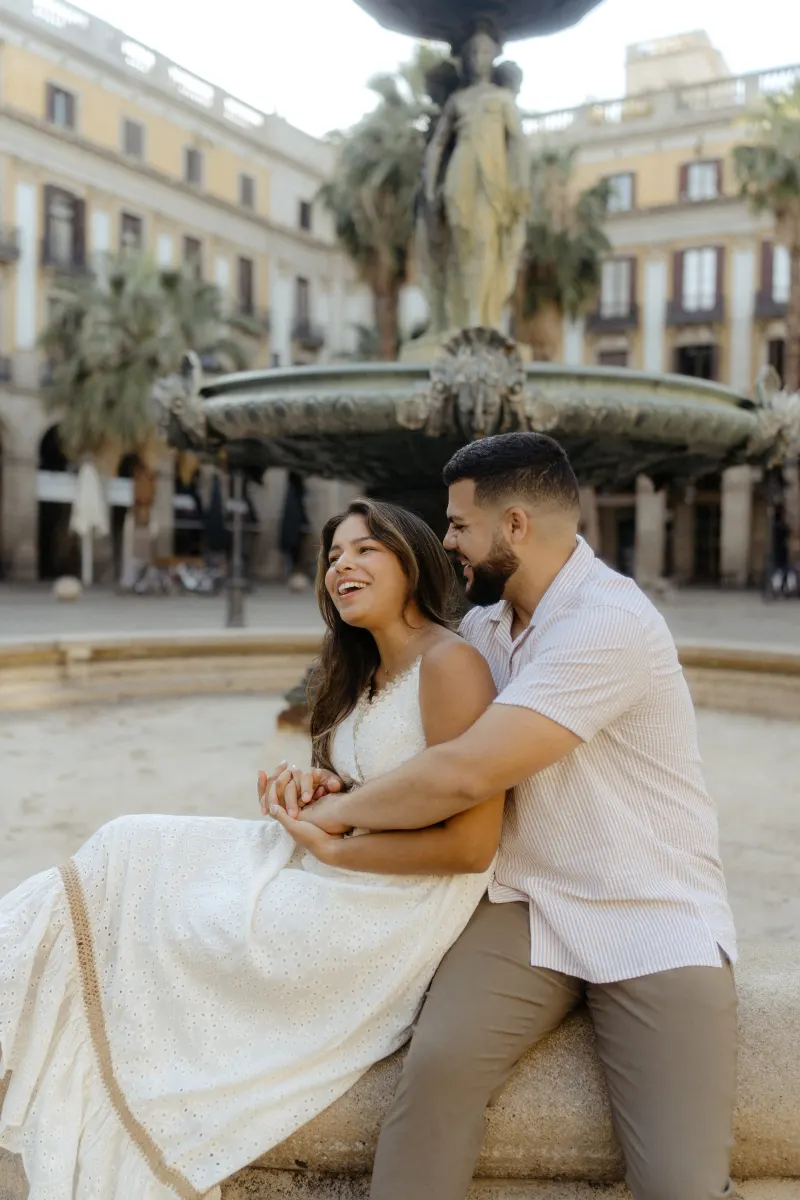 Couple laughs together while sitting by a historic fountain during a couples & engagements photoshoot in the city plaza.