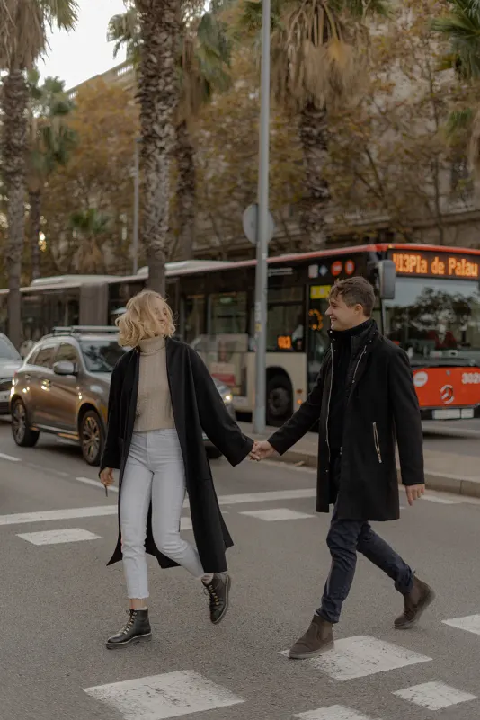 Couple laughs while crossing a city street hand in hand during a couples & engagements photoshoot in Barcelona.