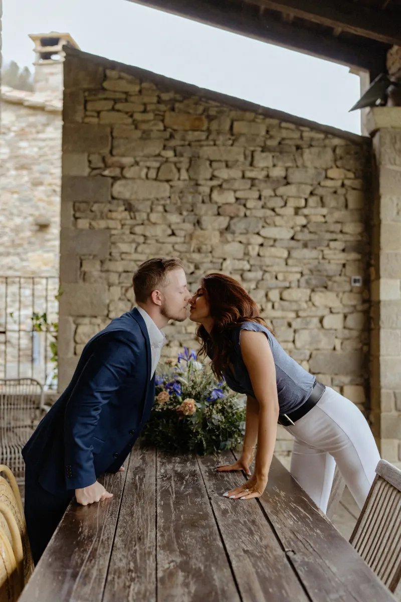 Couple leans across a rustic wooden table for a sweet kiss, celebrating their surprise proposal in the countryside.